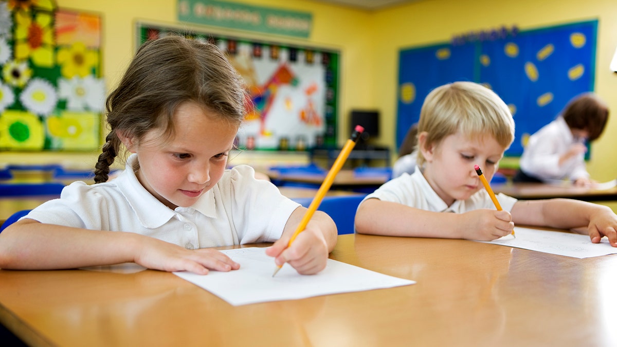 A girl and a young child write to their classroom in their desks. The little girl is writing with her left hand.