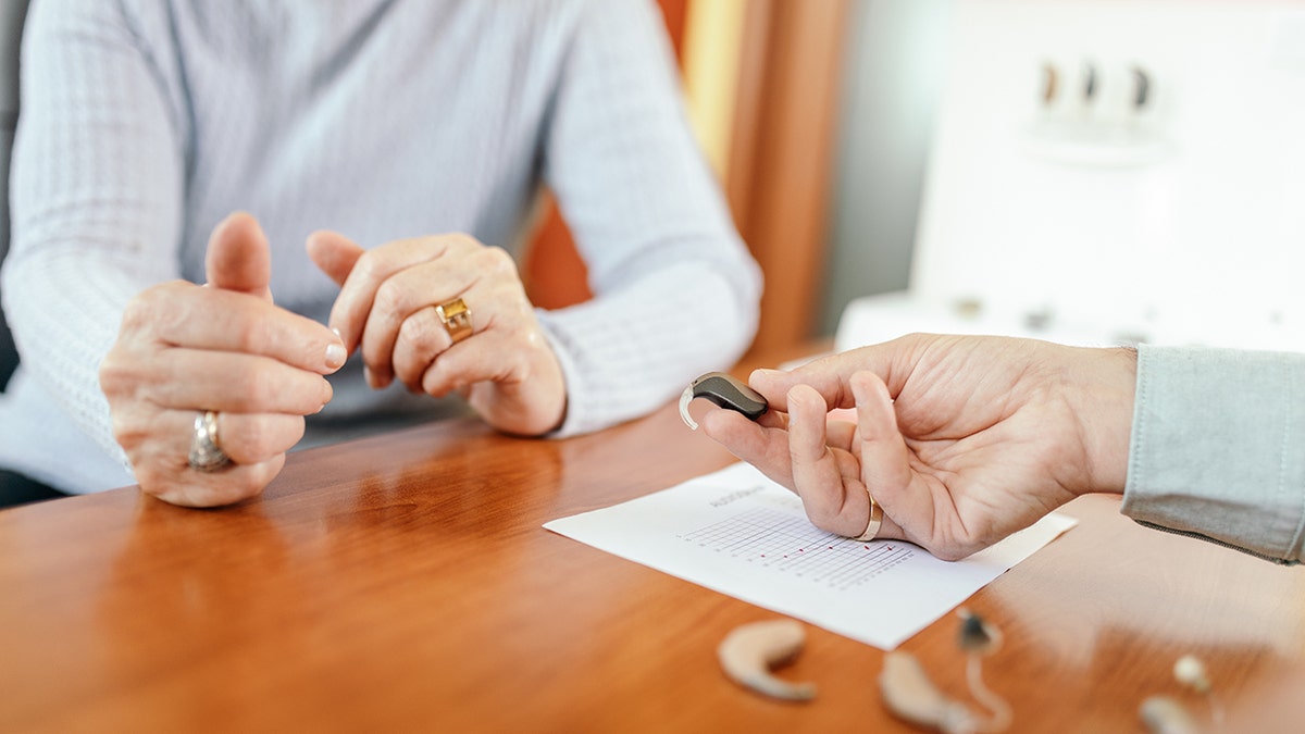 Senior woman who chooses a hearing aid device at the specialized medical clinic. Doctor who shows different types of devices. The results of the medical tests are on the counter in front. A study shows that repairing hearing loss can improve senior loneliness